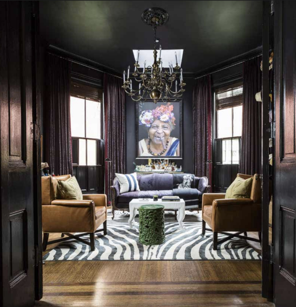 Eclectic living room with dark walls, large colorful portrait, chandelier, striped rug, tan armchairs, and settee. A small green table and a bar setup are present.