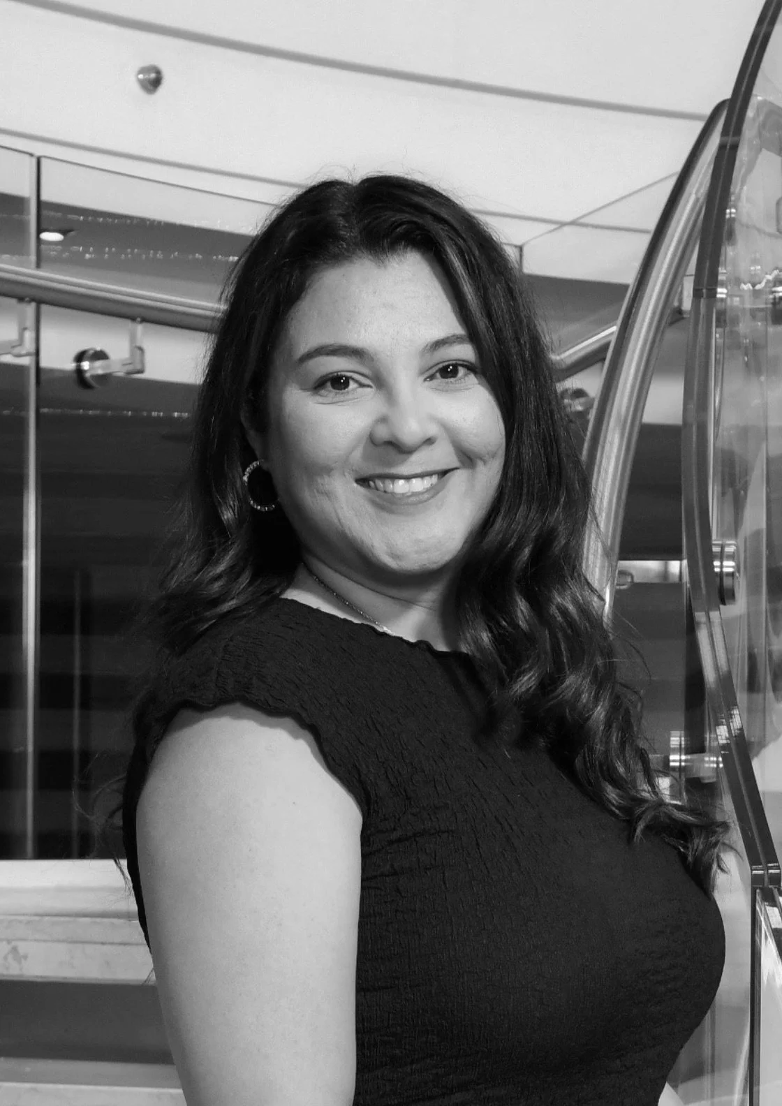 Black and white photo of a woman with wavy dark hair, smiling, wearing hoop earrings and a short-sleeved top, standing indoors near a staircase.