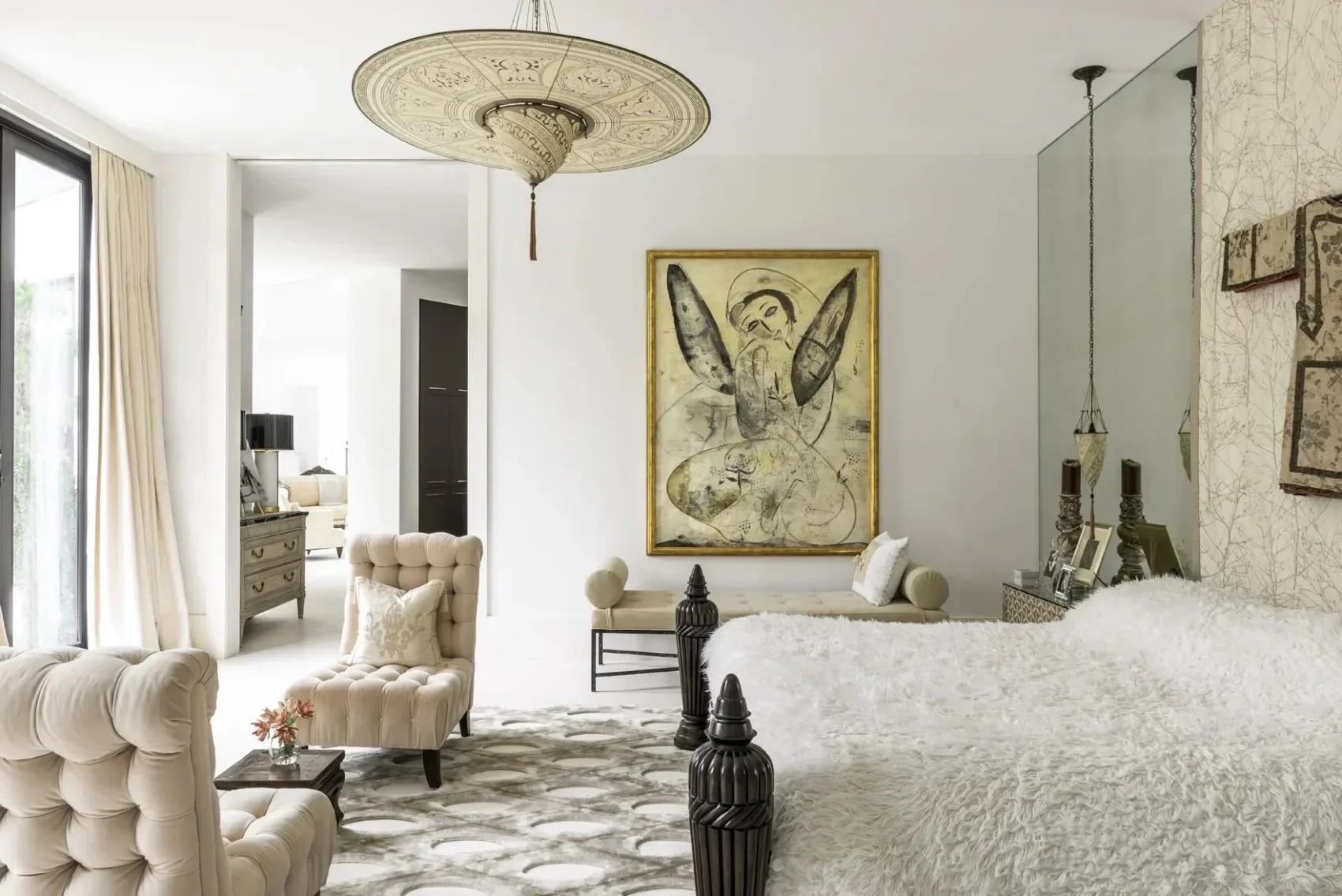 Elegant living room with cream-colored tufted armchairs and a bed with a plush white cover, a large abstract artwork of a human figure with wings on the wall, and a decorative ceiling light fixture.