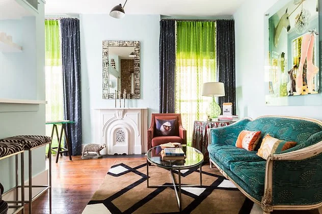 Eclectic living room with teal sofa, patterned area rug, and mixed furniture styles. Features large mirror, fireplace, glass coffee table, and decorative elements. Bright green curtains add contrast to the space.