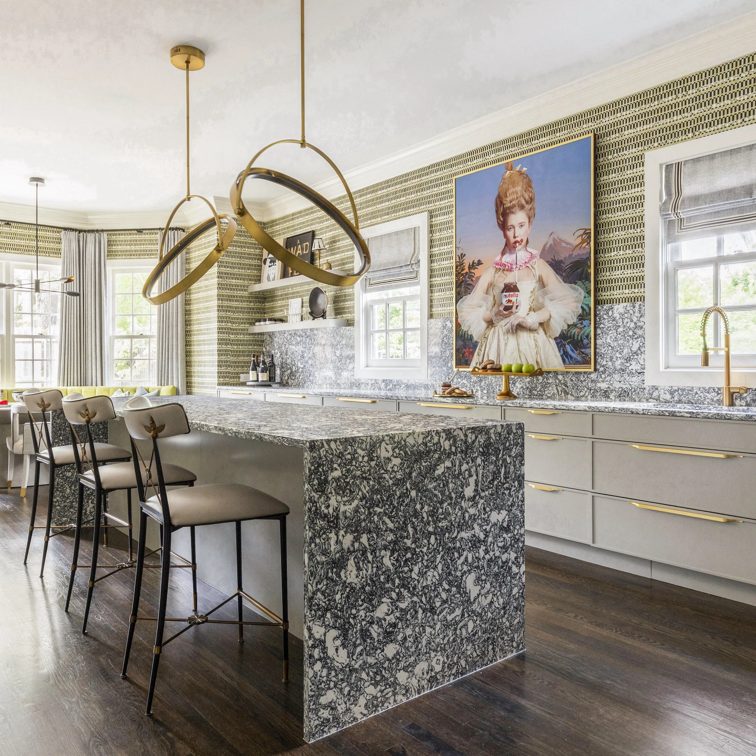 A modern kitchen with a black and white patterned granite island, white cabinets with gold handles, a large colorful portrait on the wall, large windows with light curtains, dark hardwood floors, gold pendant lighting, and various kitchen accessories.