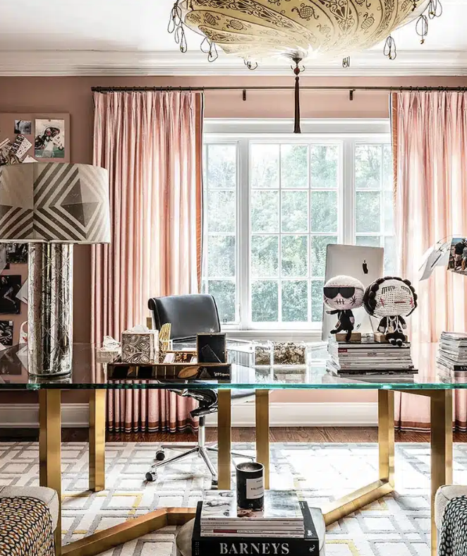 Stylish home office with glass desk, gold legs, black leather chair, decorative lamp, plush toys, books, pink curtains, and patterned ceiling light.