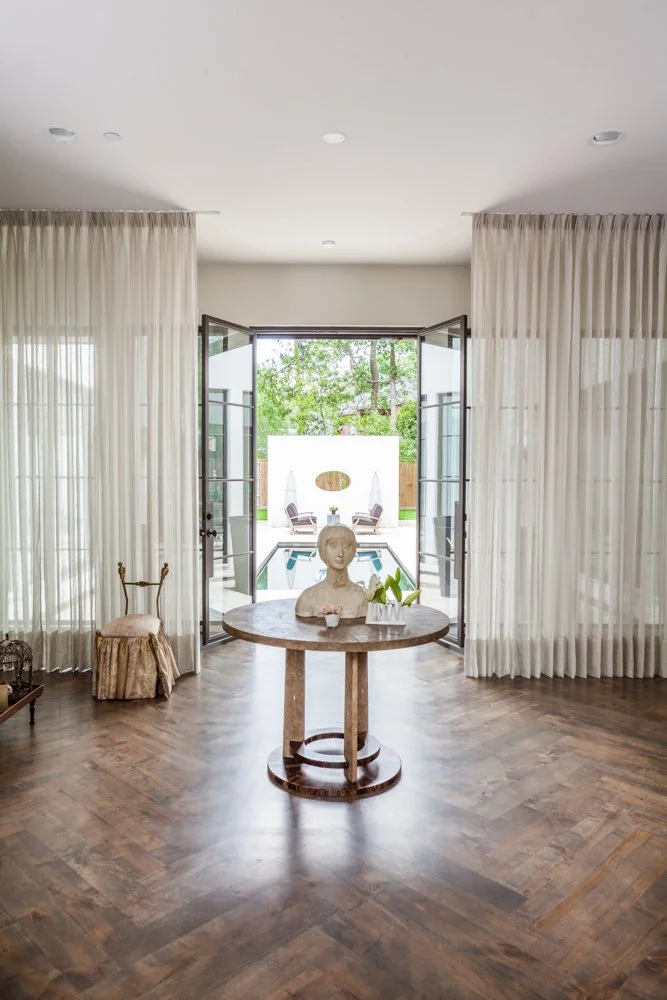 Living room with open glass doors leading to a backyard pool area, featuring a central round table with a bust sculpture and potted plants, sheer curtains, wooden floor, and backyard seating.