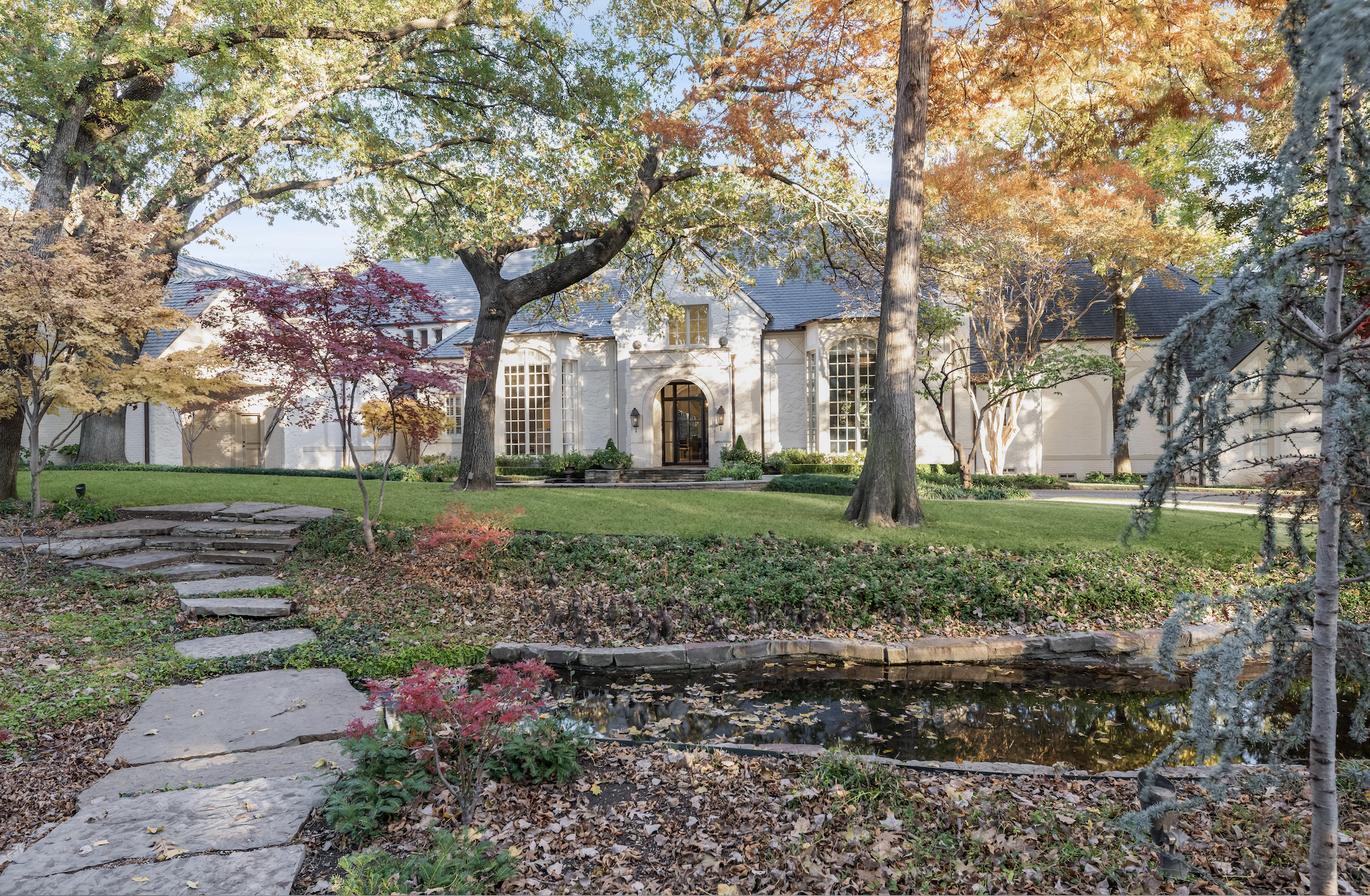 Elegant white house with large windows, surrounded by lush green lawn and a variety of trees in autumn colors. Stone path and small pond in the foreground.
