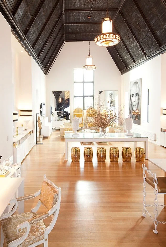 Stylish interior with a high vaulted ceiling, wooden floors, white walls, modern furniture, and large artwork. There are decorative elements like a table with gold stools and large vases, and a small chandelier hanging from the ceiling.