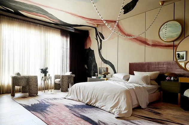 Modern bedroom with artistic wall and ceiling design, featuring a double bed with white bedding, two armchairs, a large window with sheer curtains, stylish lighting, and decorative mirrors.
