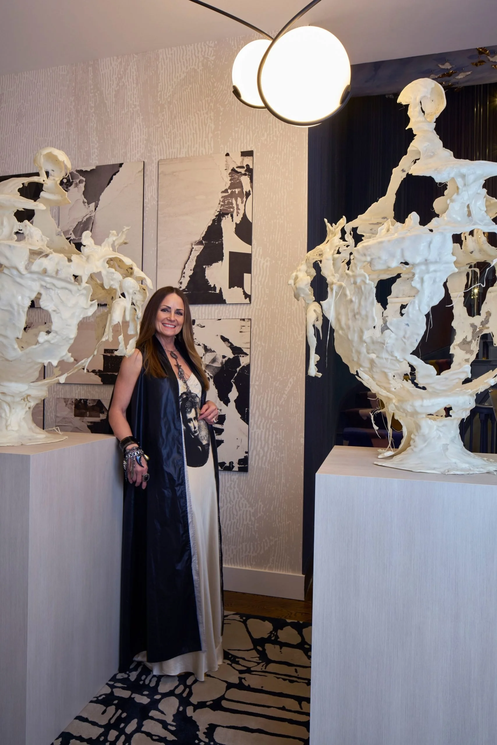 A woman standing between two abstract white sculptures on pedestals, with modern art on the wall and a spherical ceiling light above. She is wearing a long dress with a vest and layered necklaces.
