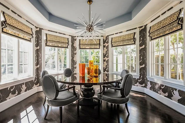 Elegant dining room with a round glass table, modern chairs, and a starburst chandelier, surrounded by large windows with patterned shades and dark hardwood flooring.
