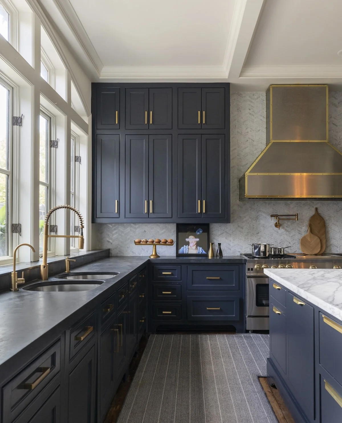 Modern kitchen with navy blue cabinets, gold hardware, marble countertops, large windows, and a stainless steel range hood.