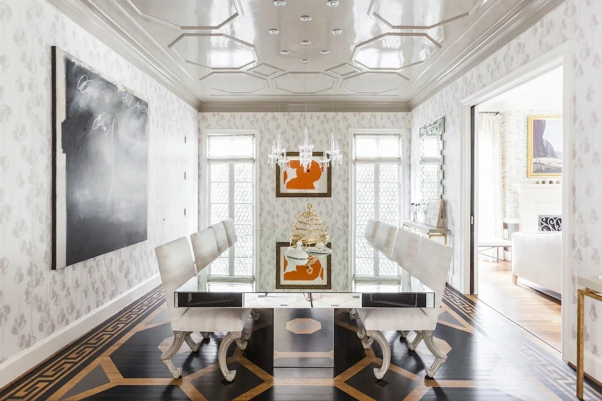 Elegant dining room with a mirrored table, white upholstered chairs, chandelier, wall art, and patterned floor mats, with sunlight streaming through windows.