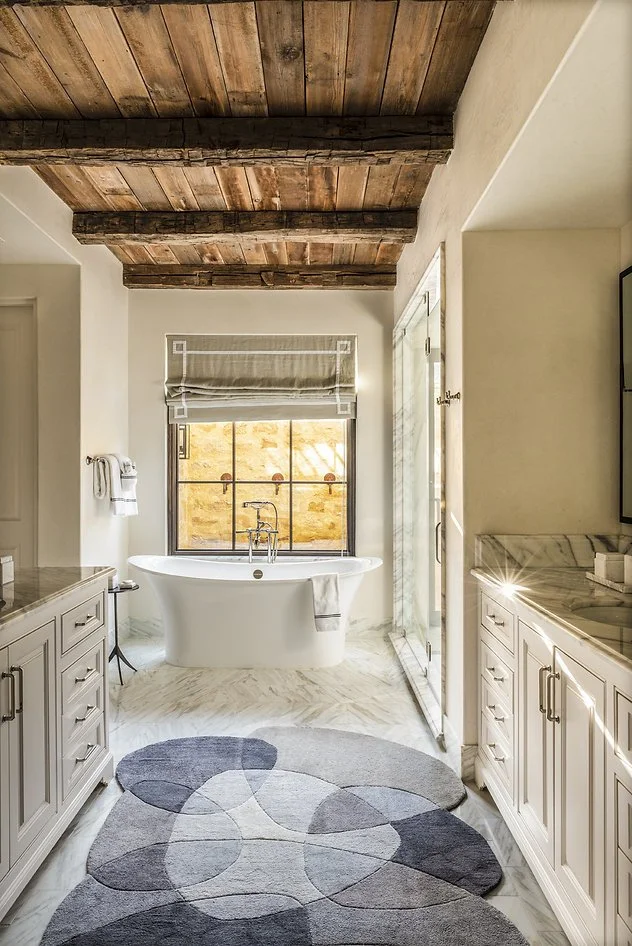 A luxurious bathroom with a rustic wooden ceiling, a freestanding white bathtub near a large window, a patterned area rug on the marble floor, and white cabinetry on both sides.