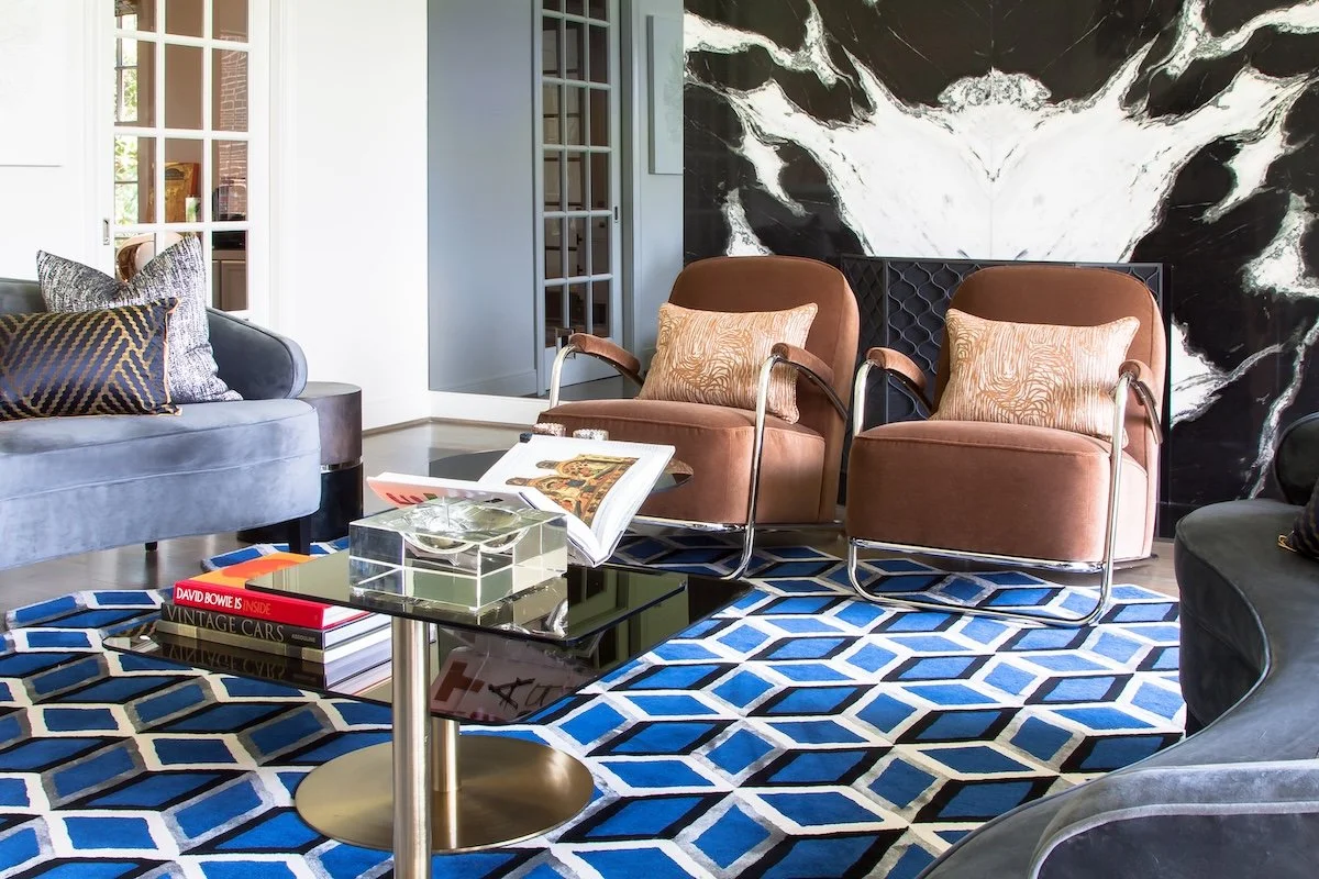 A modern living room featuring two brown cushioned armchairs with patterned pillows, a velvet gray sofa with decorative pillows, a glass-top coffee table with books and a decorative bowl, a vibrant blue and white geometric area rug, and a black and white marble wall art piece in the background.
