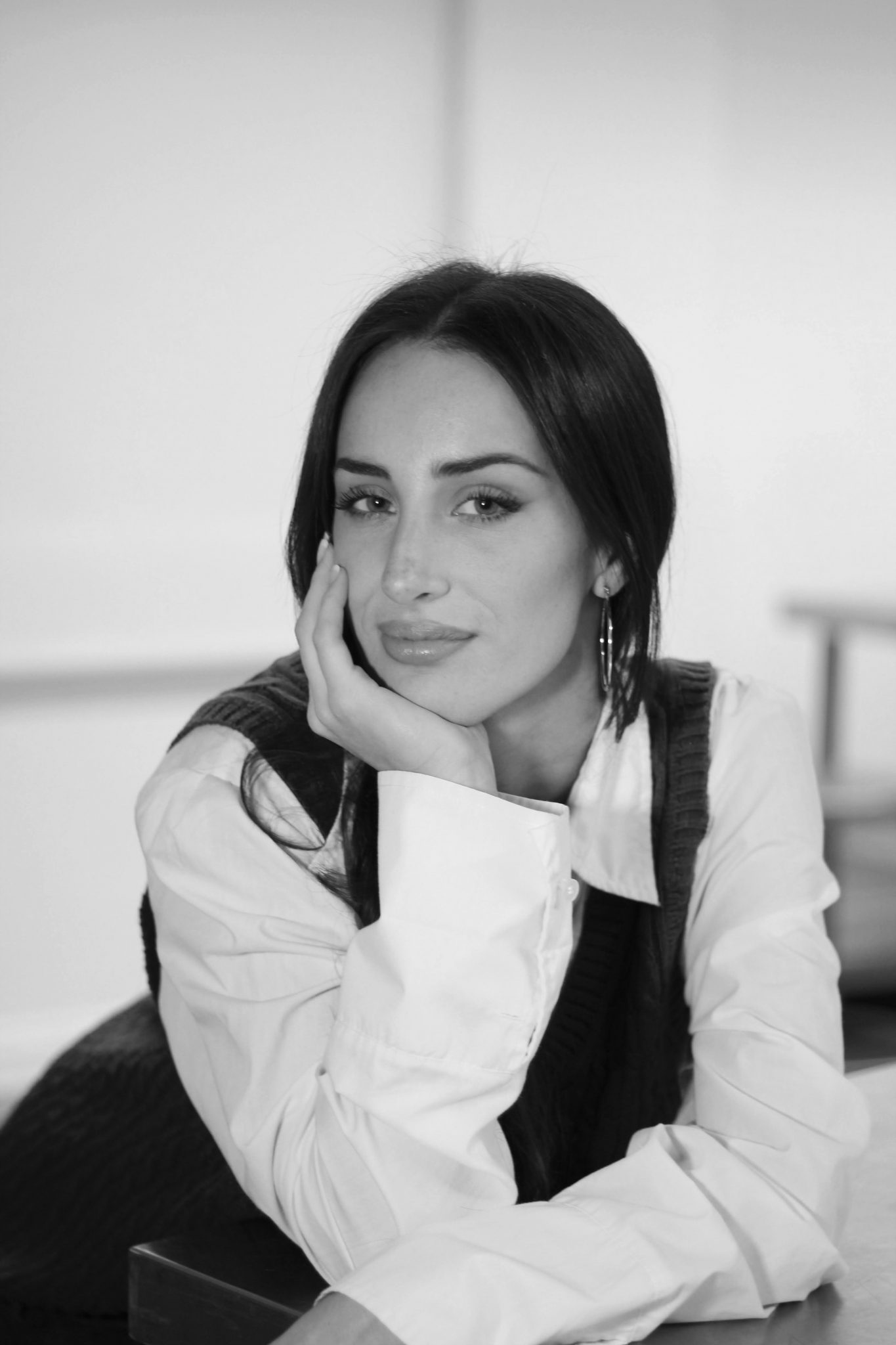 Black and white portrait of a woman with long dark hair, resting her chin on her hand, wearing a white shirt and a vest, looking at the camera.
