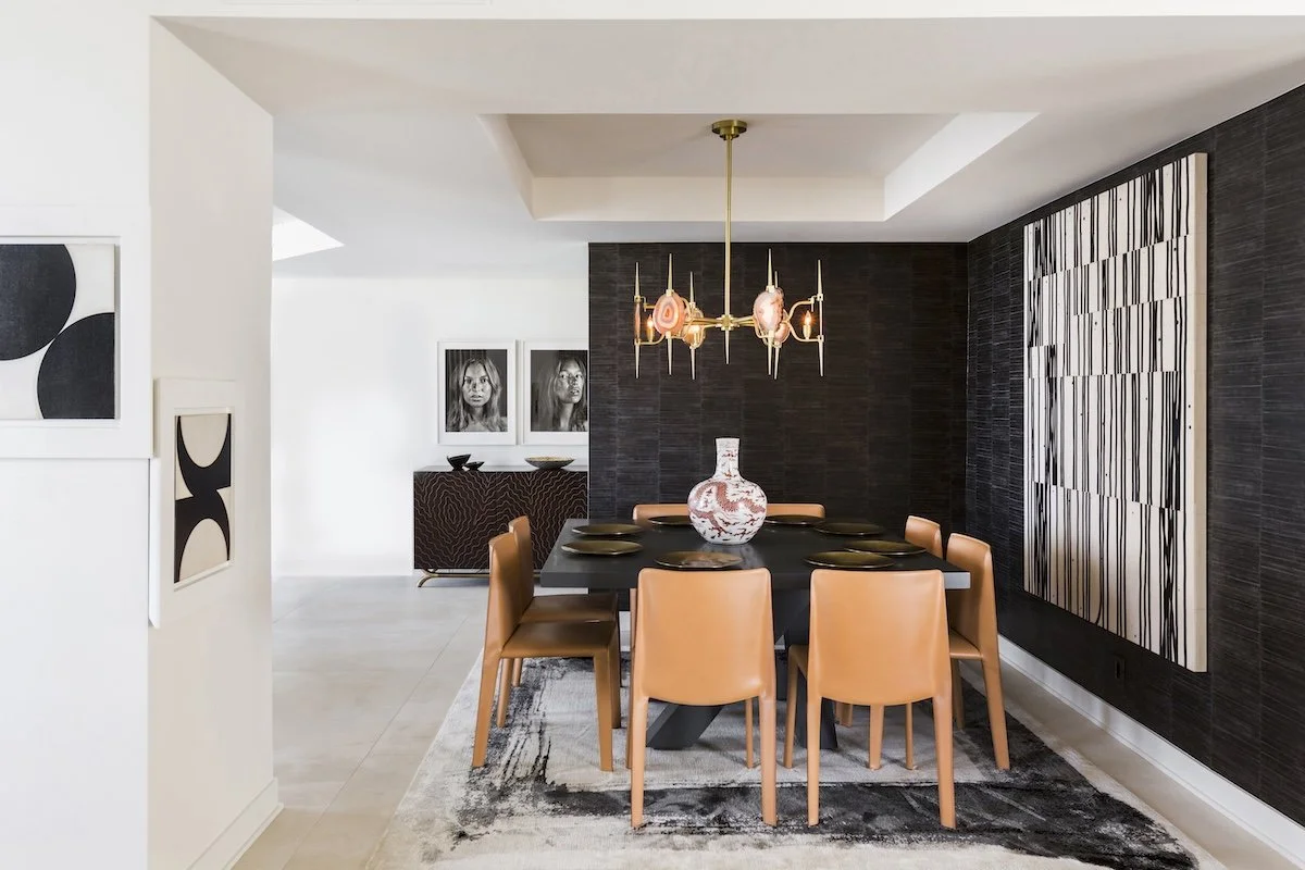 Modern dining room with six tan chairs around a black table, decorative vases on the table, and a chandelier hanging above. Artworks on the walls include black and white portraits and abstract designs.