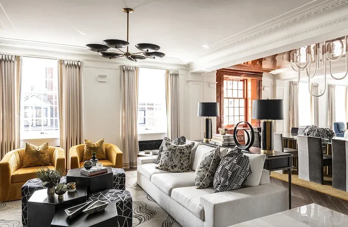 Spacious living room with yellow armchairs, a white sofa with patterned cushions, black and white coffee tables, large windows with curtains, modern chandelier, and a dining area with gray chairs and black lamps.