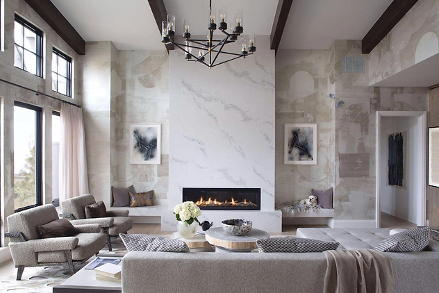 Modern living room with high ceiling, decorative wall panels, and a central fireplace. The room features contemporary furniture, including sofas and armchairs, accented by a black chandelier. Large windows provide natural light and decorative art pieces are hung on the walls.