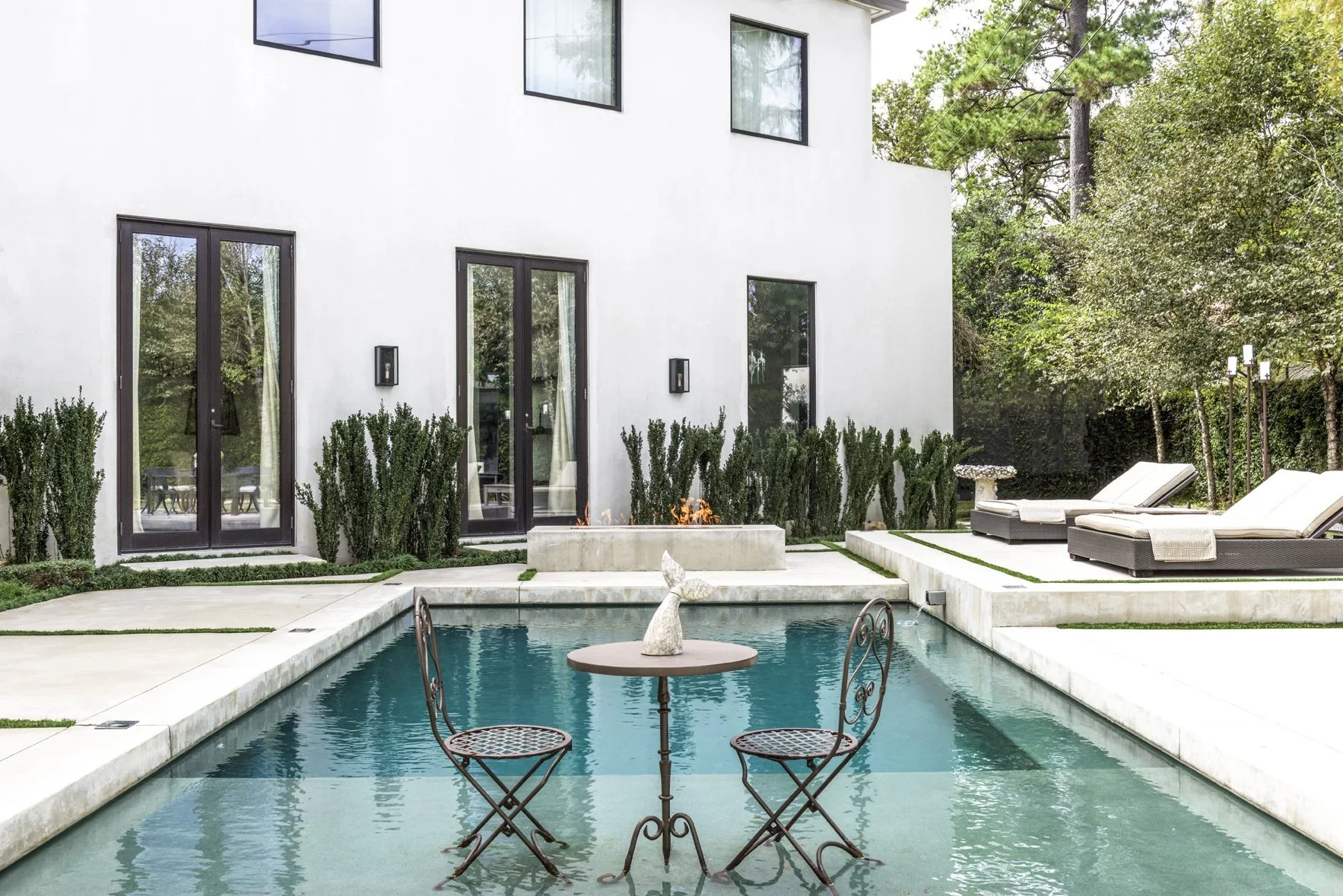 A backyard with a swimming pool, a small round table with two chairs, lounge chairs, and a modern white house with large windows. There are bushes, trees, and outdoor lighting.