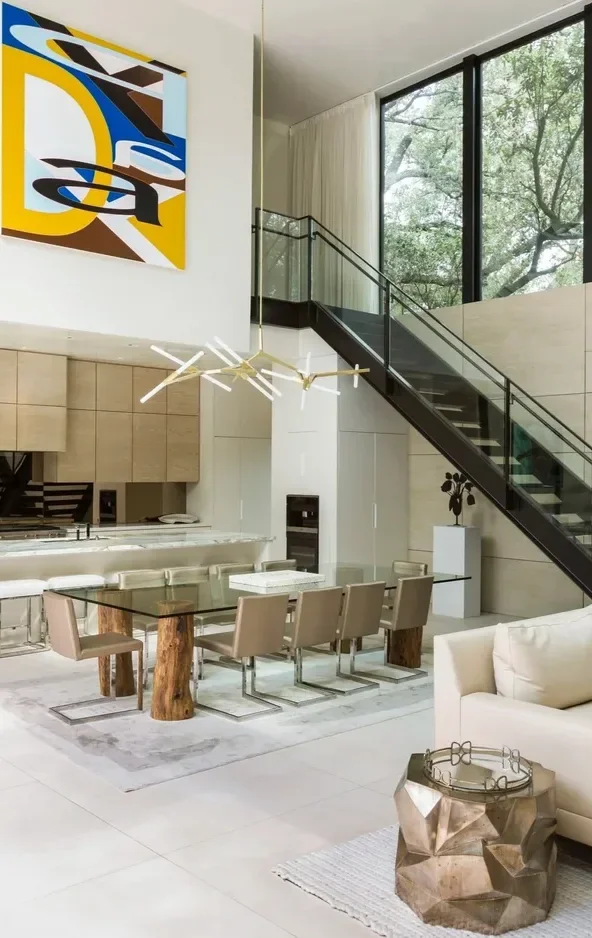 Modern dining area in a spacious, bright living room with a glass dining table, beige chairs, a contemporary chandelier, a white sofa, and large windows showing trees outside.