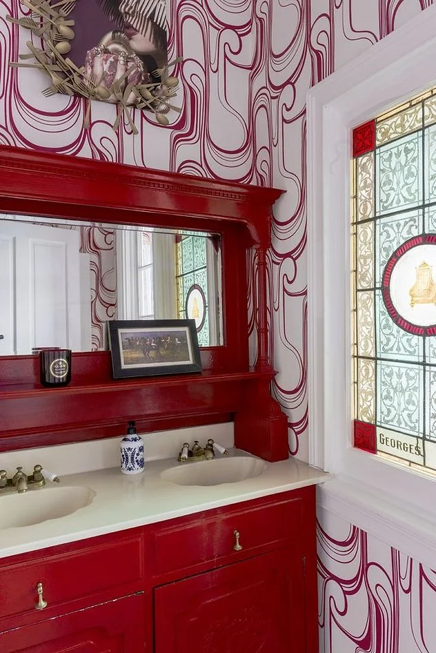 Bathroom with red vanity, double sinks, decorative wall mirror, unique swirling wallpaper, stained glass window, and framed artwork.