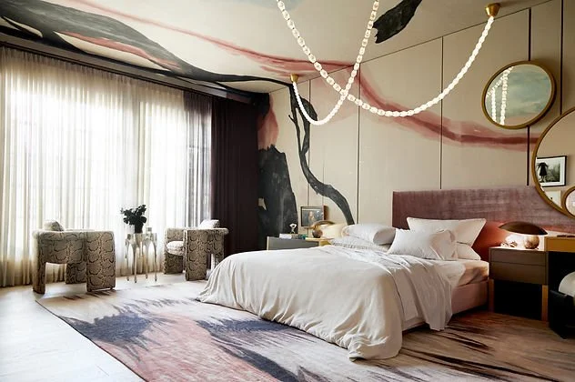 Modern bedroom with abstract wall art, large windows with sheer curtains, a bed with white linens, upholstered chairs, a geometric rug, and a decorative ceiling light.