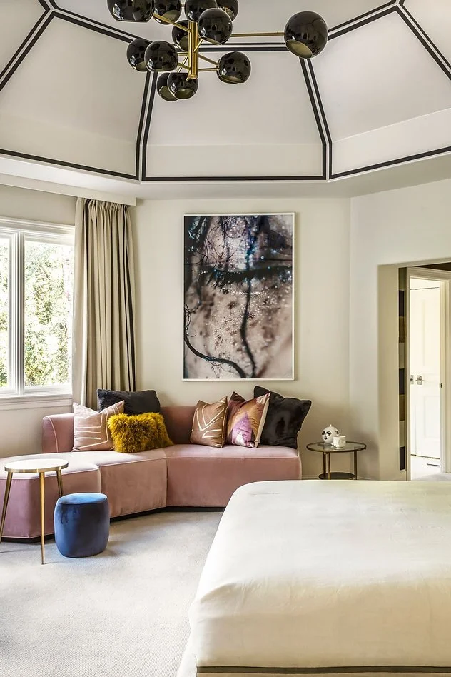 Modern bedroom interior with a pink sectional sofa, colorful pillows, round tables, and a bed. A large abstract artwork is on the wall. The room features a unique geometric ceiling with a black orb chandelier.
