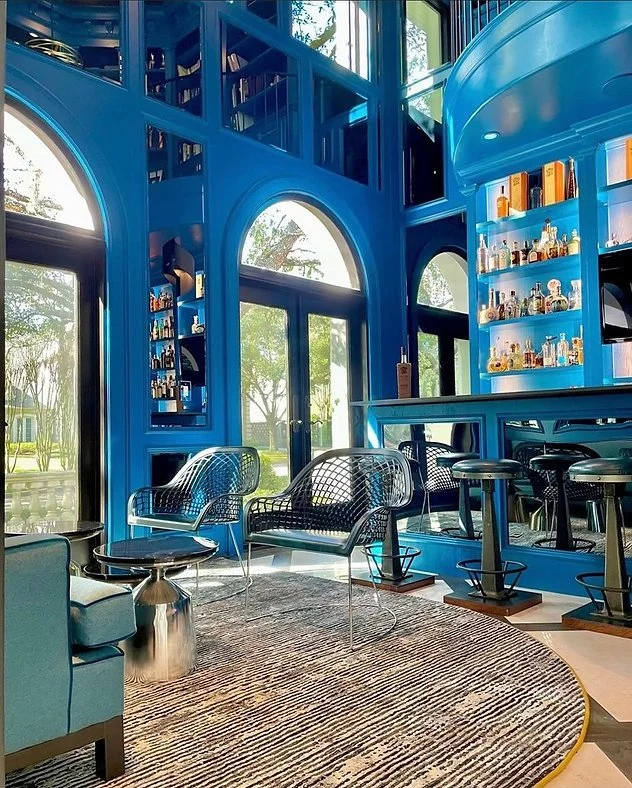 A modern home bar with a blue interior, including a bar counter stocked with bottles, high stools, and metal chairs. Large windows and mirrors surround the space, creating a spacious atmosphere. A round rug and small side table complete the decor.