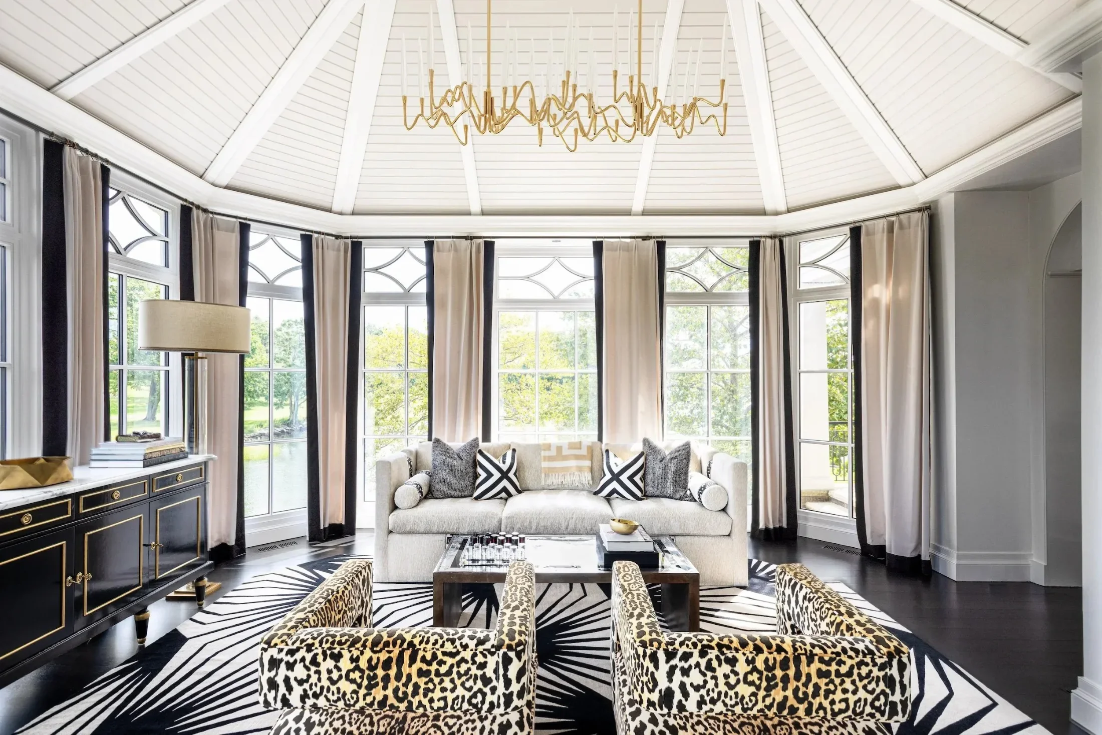 Elegant living room with large bay windows, beige and black curtains, patterned armchairs with leopard print, white sofa with black and white pillows, black and gold sideboard, zebra print rug, and a gold abstract chandelier.