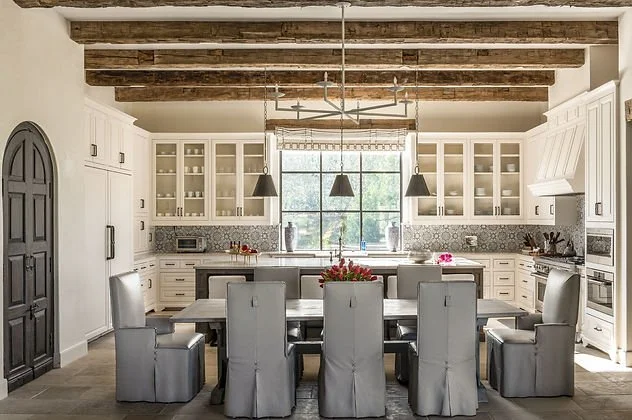 Rustic modern kitchen with wooden beams, large dining table with eight chairs, pendant lights, and white cabinetry.