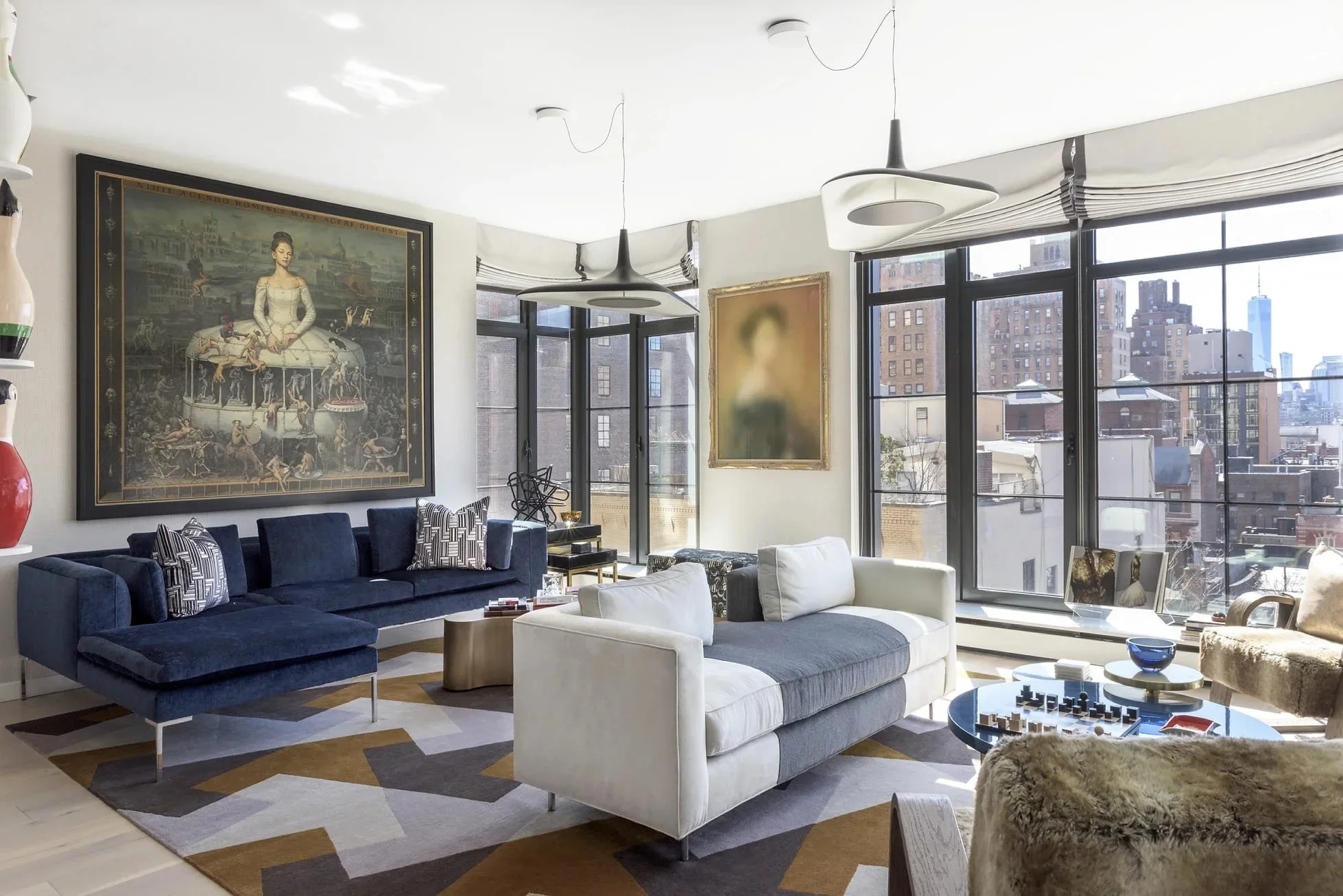 A modern living room with large windows showing a city skyline. The room has a navy blue sectional sofa with patterned pillows, a white armchair, and a patterned rug. Large framed artwork hangs on the wall, and there are contemporary pendant lights hanging from the ceiling.