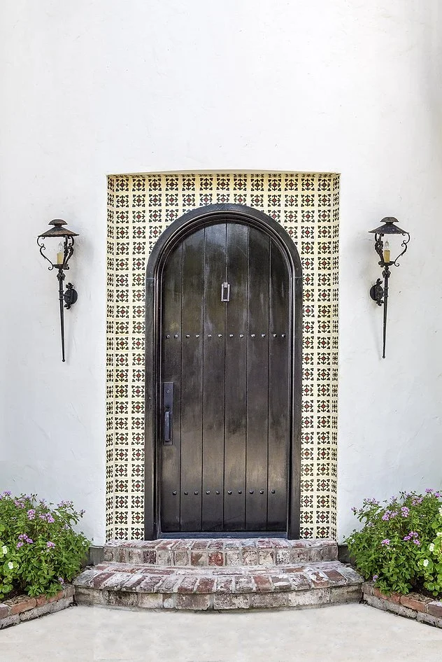 Arched wooden door with decorative tile border, flanked by two wall-mounted lanterns, surrounded by stucco wall and brick steps, with potted plants.