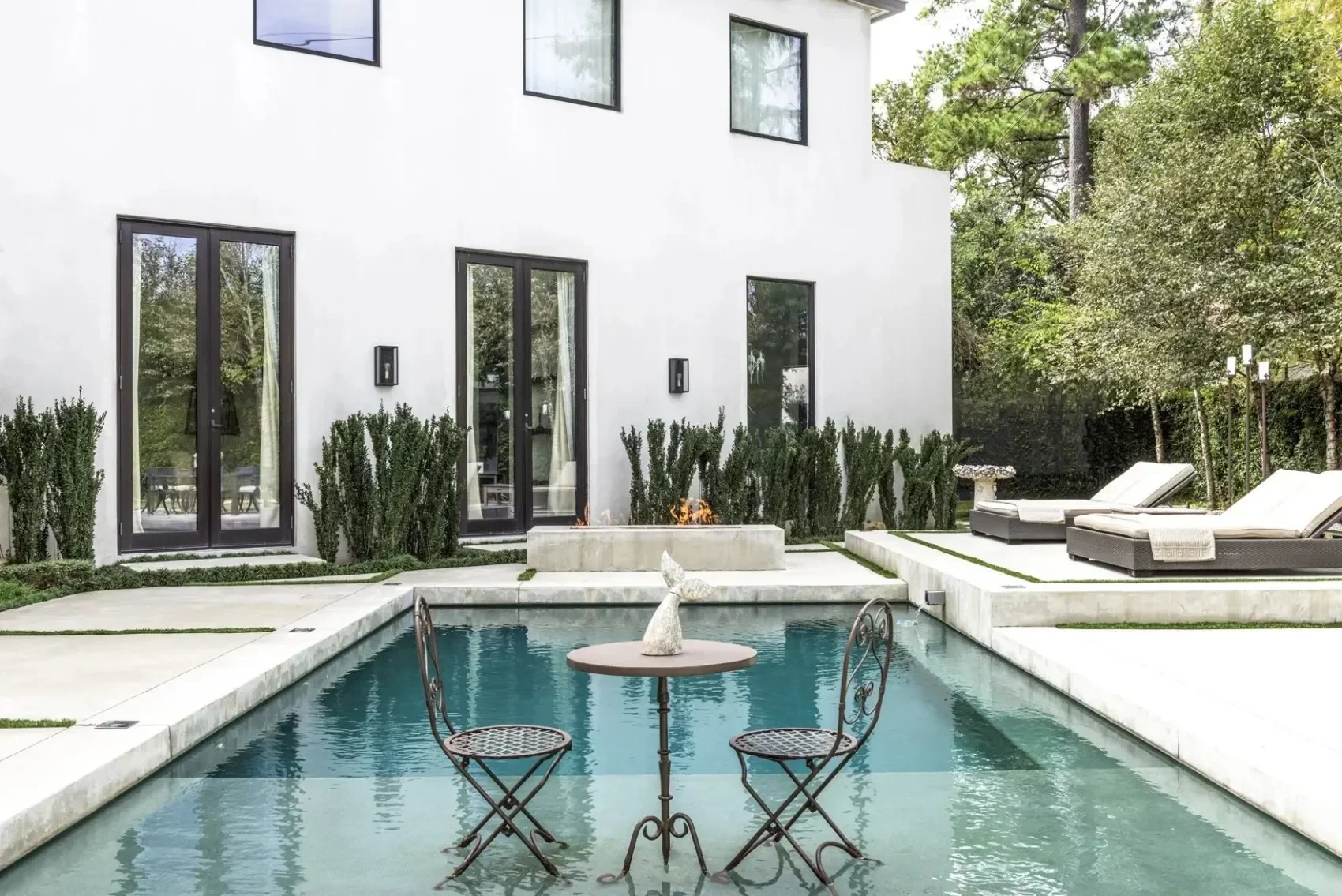 Modern white house with large glass doors and windows, outdoor swimming pool with a rooster-shaped fountain, patio furniture, and greenery surrounding the backyard.