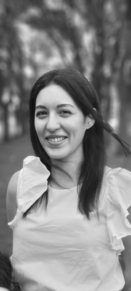 Black and white photo of a woman smiling outdoors, with blurred trees in the background.