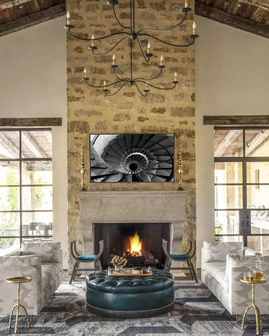 Living room with stone fireplace, two white sofas, two chairs with blue cushions, a black leather ottoman with tray, chandelier, and a large flat-screen TV above the fireplace, with large windows on each side.