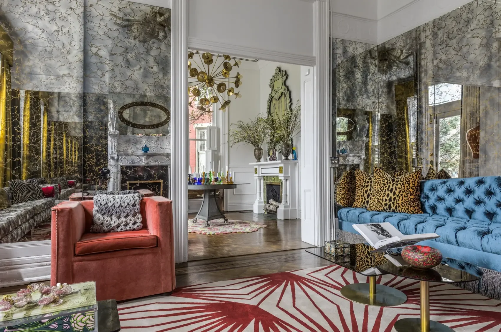 Well-decorated living room with mirrored walls, various sofas with patterned and animal print pillows, a glass coffee table with books and decorative objects, a fireplace, and a view into a neighboring room with a chandelier, vases, and large windows.