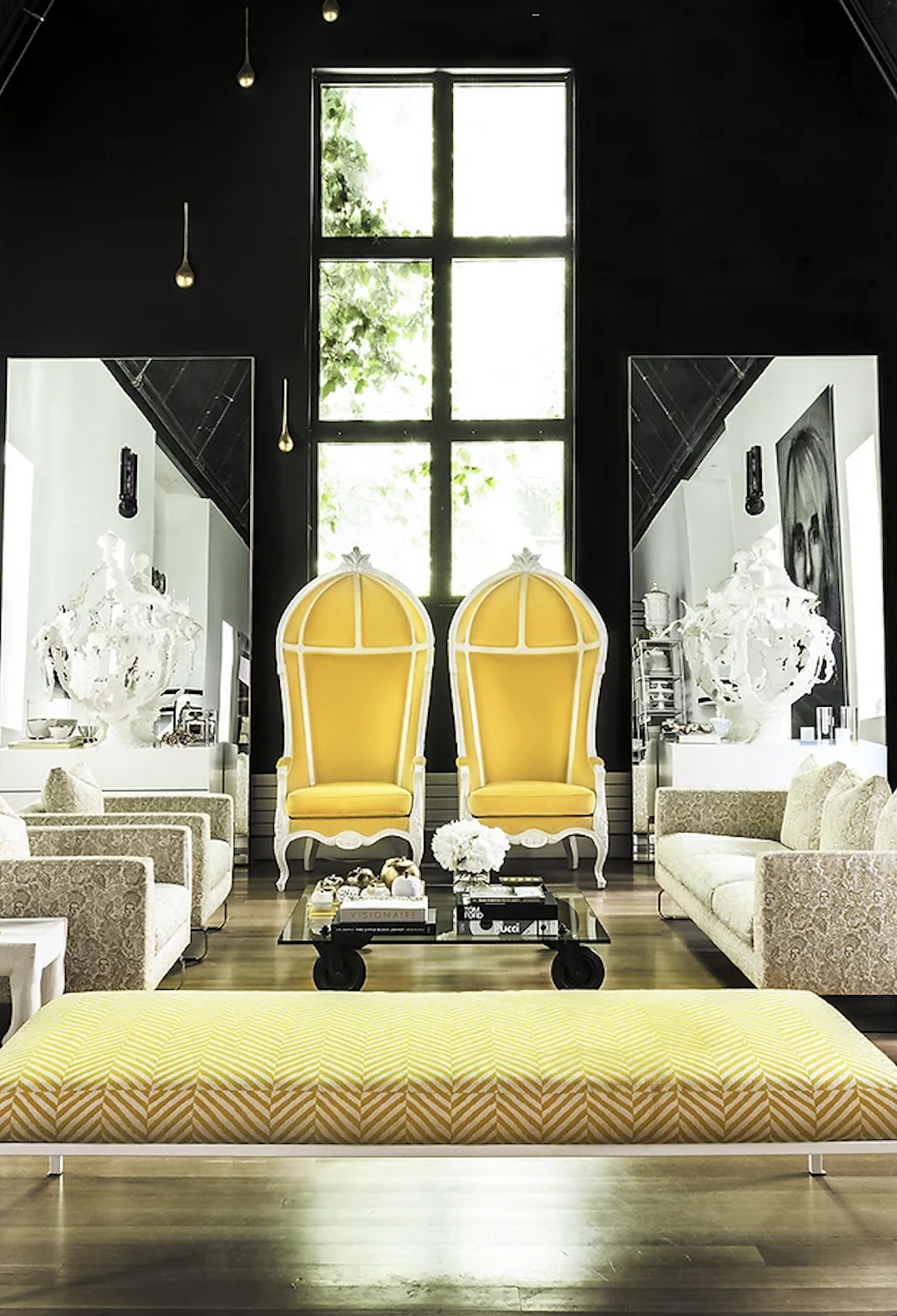 Modern living room with yellow accent chairs, mirrored walls, a large window, contemporary artwork, neutral sofas, and a glass coffee table with white flowers.