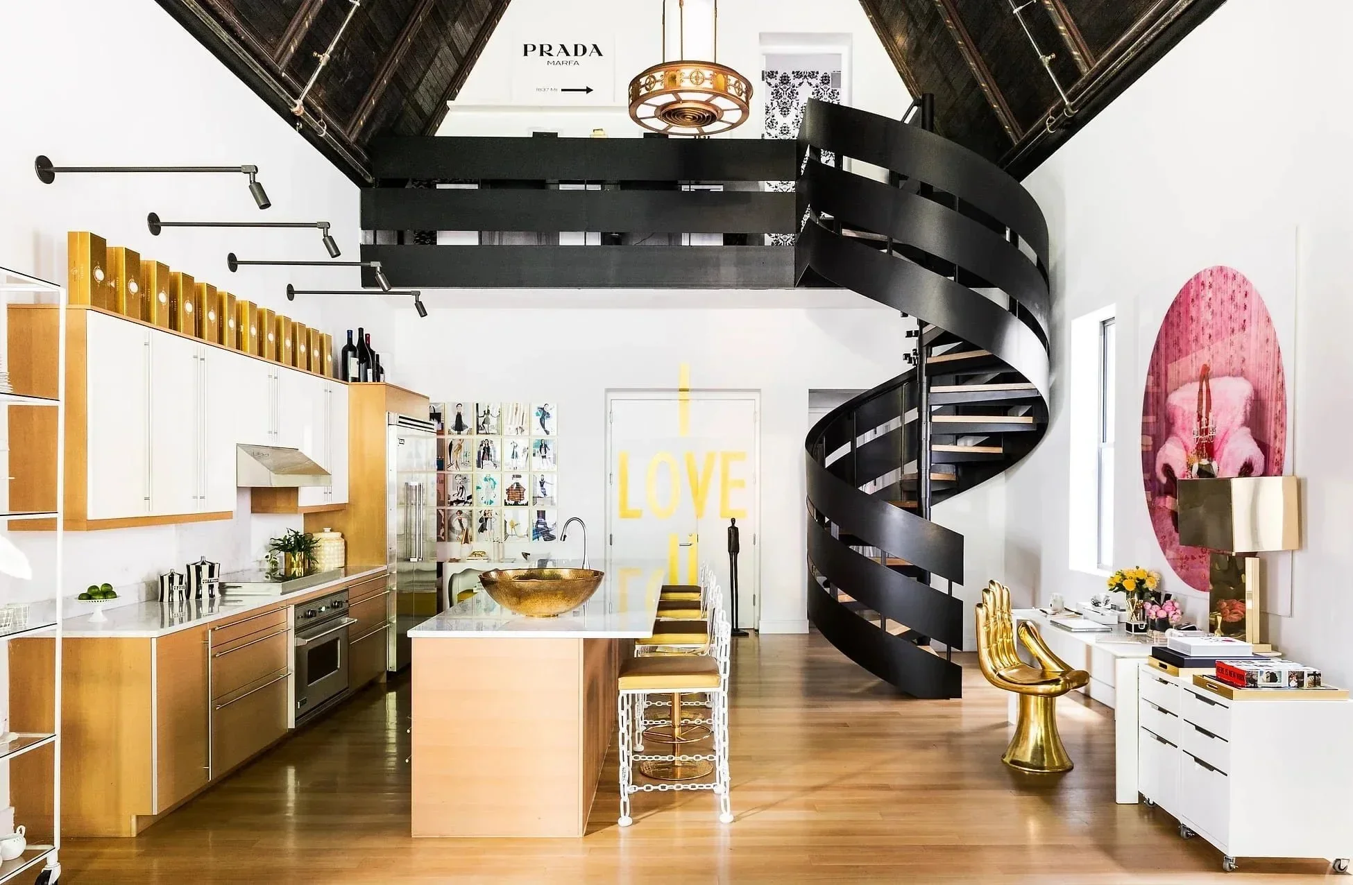 Interior of a modern, stylish kitchen and living space with a black spiral staircase, white and wooden cabinets, colorful artwork, and decorative objects including gold accents, bottles, and flowers.