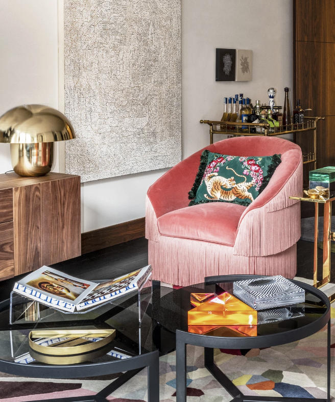 Modern living room with pink velvet chair, tiger pillow, brass lamp, abstract art, coffee table with books, and a bar cart with bottles.