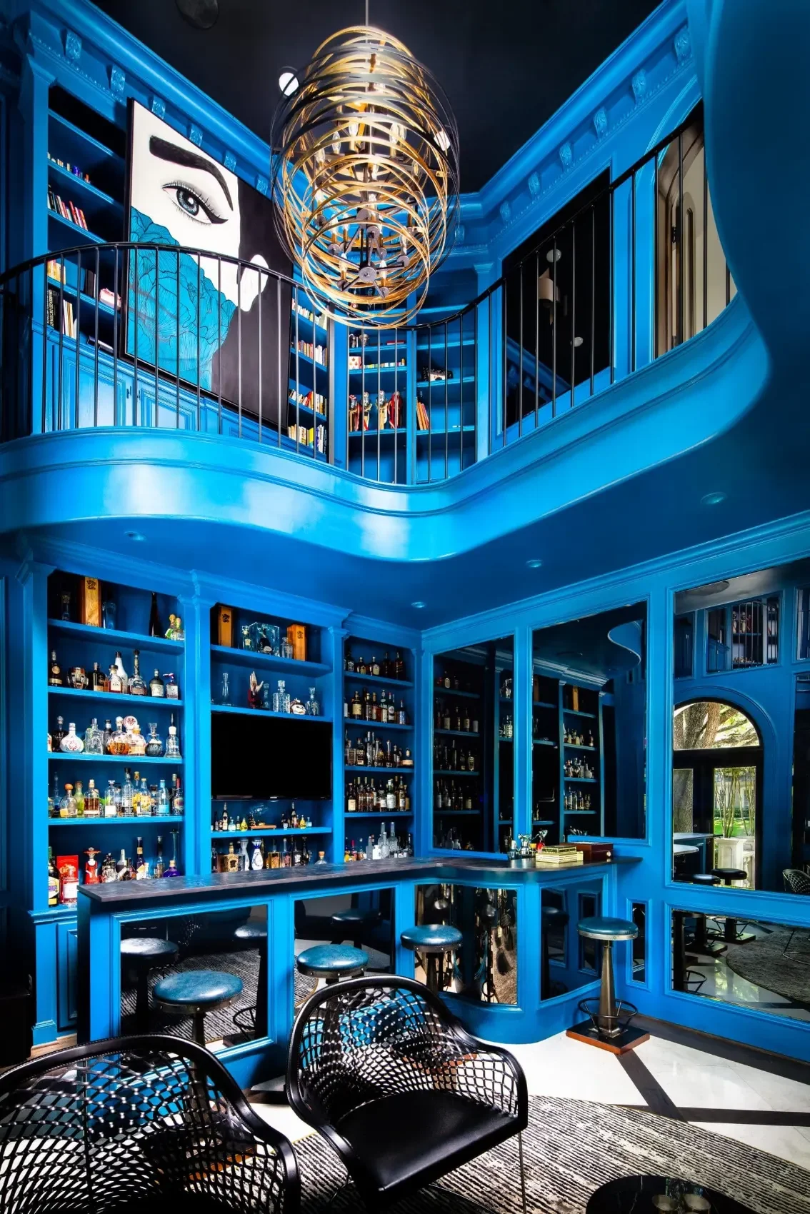 A vibrant blue bar interior with tall shelves of liquor bottles, a seating area, a bar counter, and a spiral chandelier hanging from the ceiling, accented by a large artwork of a woman's eye on the wall.