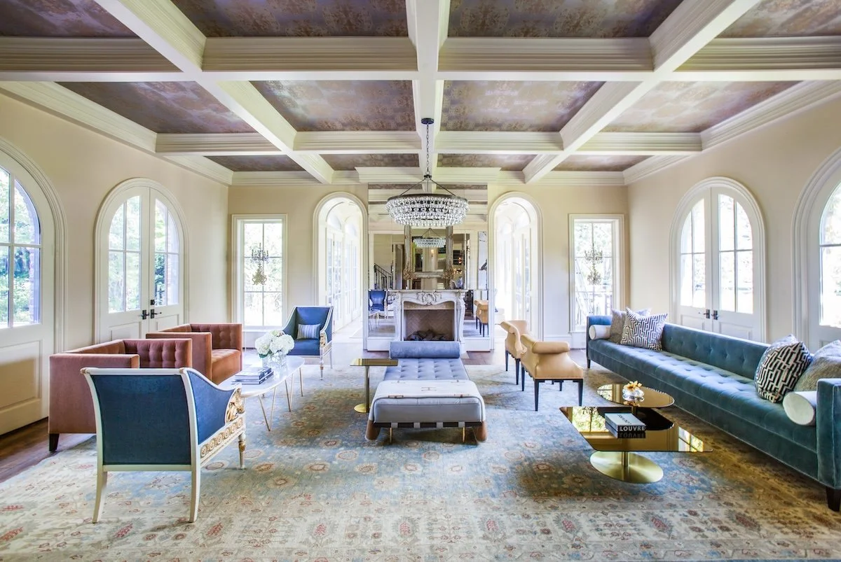 Elegant living room with large windows, a coffered ceiling, a central chandelier, a fireplace, and various colorful sofas and armchairs arranged around a coffee table, with a decorative rug on the wooden floor.