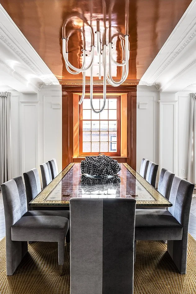 Modern dining room with a large table, gray chairs, geometric black centerpiece, and contemporary chandelier. Ceiling is reflective copper-colored, and there is a window with an orange frame.