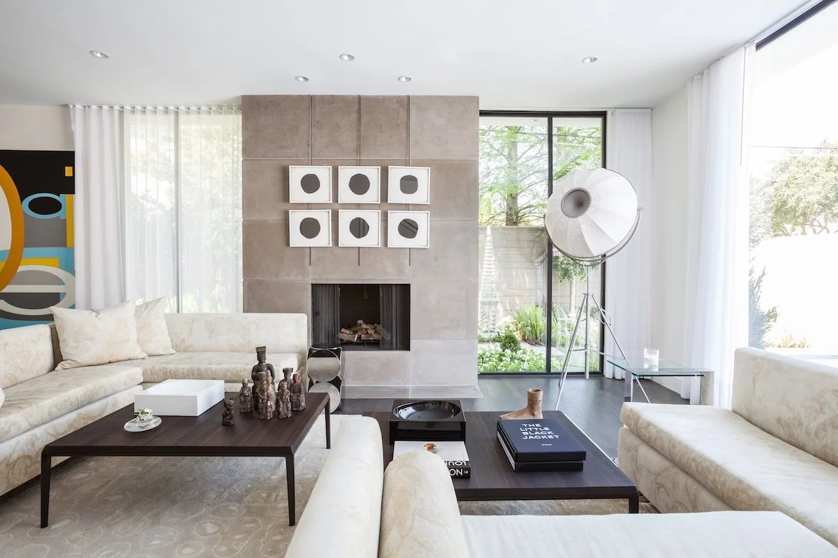 Modern living room with white sofas, a dark wood coffee table, a large floor lamp, and a fireplace set into a gray tiled wall, with large window and sliding glass door showing outdoor greenery.
