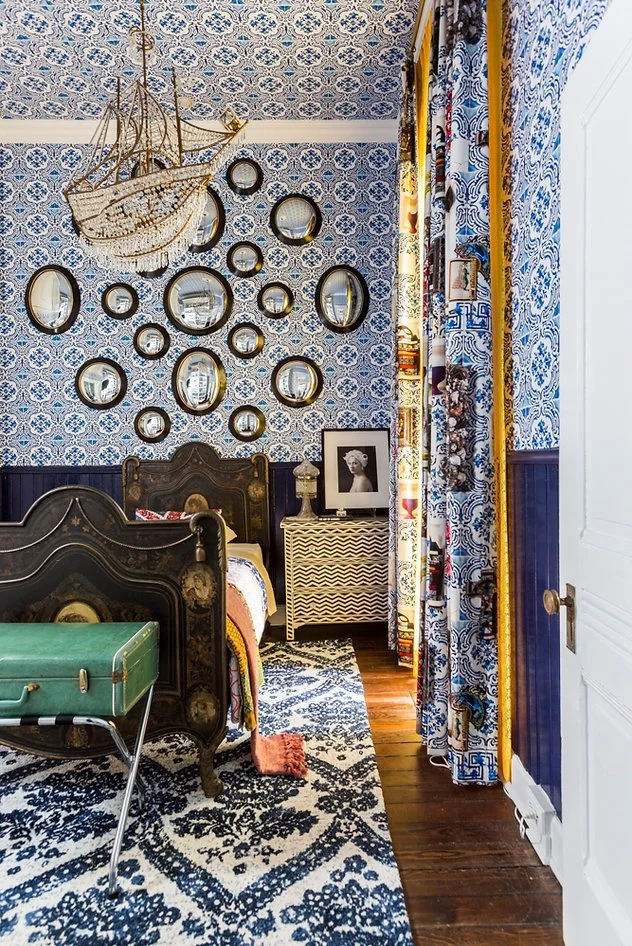 Room with blue patterned wallpaper, multiple round mirrors, ornate black bed, green suitcase, chandelier shaped like a ship, and a patterned rug.